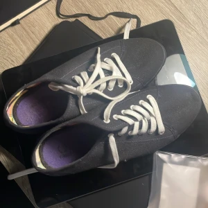 Svarta sneakers från Fred Perry - Snygga svarta sneakers från Fred Perry med vita snören och diskret logga på sidan. Skorna har en klassisk låg profil, rund tå och platt sula. Insidan har ett färgglatt rutigt foder och lila innersula. Perfekta för en clean och stilren look.