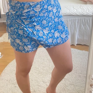 Blå blommiga omlottshorts med snörning - Snygga blå shorts med vitt blommigt mönster och omlottdesign. De har snörning i sidan och är perfekta för varma dagar. Lätta och luftiga, passar dig som gillar en avslappnad men trendig stil. Små i storlek skulle säga att detta passar S