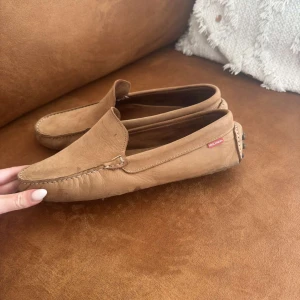 Beige mockaloafers från Wisconsin - Snygga loafers i beige mocka från Wisconsin. Skorna har klassisk design med rund tå, dekorativa sömmar och gummidetaljer på hälen för extra grepp. Perfekta för en chill och stilren look.