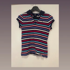 Randig pikétopp från Tommy Hilfiger - Säljer en färgglad randig pikétopp från Tommy Hilfiger med korta ärmar och klassisk krage. Toppen har marinblå, röd, rosa, vit, blå och lila ränder samt liten logga på bröstet.