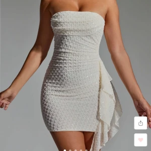 Vit glittrig kort klänning med volang - Superfin vit kort klänning med glittriga detaljer och volang på sidan. Axelbandslös med öppen rygg och två tunna band bak. Tight passform och strukturerat tyg som ger en lyxig känsla. Perfekt för dig som vill sticka ut och glänsa. Man kan fixa banden där back, om man vill att det ska va tajtare eller lite större, där back på klänningen. Har även använt klänningen 1 gång