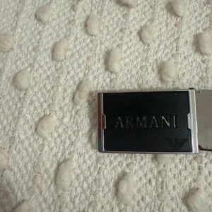 Svart skärp från Armani i skinn - Snyggt svart bälte från Armani med stilren, rektangulär metallspänne där loggan och namnet är ingraverat. Bältet är gjort i äkta skinn och har en klassisk design som passar till jeans eller kostymbyxor.