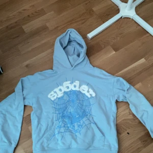 Ljusblå Sp5der hoodie med tryck - Säljer en ljusblå hoodie från Sp5der med stort spindelnäts-tryck och 3D-liknande vit text på bröstet. Tröjan har huva och är tillverkad i 100% bomull, perfekt för dig som gillar streetwear och unika prints.