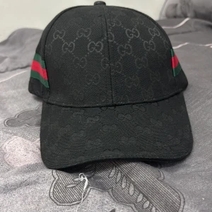 Svart Gucci keps med GG-mönster - Svart keps från Gucci med klassiskt GG-monogram över hela ytan och gröna/röda ränder på sidorna. Justerbar rem baktill i läder och insida med Gucci-logga på banden. Snygg och ikonisk accessoar för dig som gillar streetstyle och lyx.