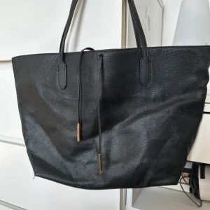Svart väska i skinnimitation - Stor svart shoppingväska från H&M i skinnimitation med två långa handtag och dekorativa snören med metalländar. Väskan har ett rymligt fack och en innerficka med dragkedja. Perfekt för dig som vill ha plats för allt du behöver under dagen.