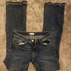 Mörkblå bootcut jeans från Lindex - Snygga mörkblå jeans från Lindex i bootcut-modell. Klassisk femficksdesign, låg midja och råa kanter nertill för en trendig look. Tillverkade i slitstarkt denim med lätt stretch för skön passform.