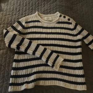 Randig stickad tröja Ralph Lauren XS - Snygg stickad tröja från Ralph Lauren i beige och svart med breda ränder. Tröjan har rund halsringning, långa ärmar och tre dekorativa knappar på ena axeln. Perfekt för dig som gillar en klassisk men chill stil.