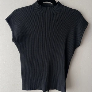 Svart ribbad topp med snöring - Säljer en svart ribbad topp med ärmlös design. Toppen har en unik detalj med snörning och rund öppning baktill. Perfekt för dig som gillar stilrena plagg med en twist. Snygg att matcha med både jeans och kjol.