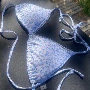 Blommig bikiniöverdel från Bik Bok - Supersöt trekantsbikini från Bik Bok med vitt tyg och småblommigt blått mönster. Knytband i nacken och ryggen för perfekt passform. Liten volangkant längs kuporna ger en extra gullig touch. Perfekt för strandhäng och soliga dagar!