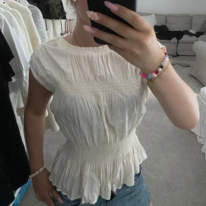 Topp - Supersöt topp från H&M med smockad midja och volangkant nertill. Blusen har korta ärmar och är gjord i ett lätt, skrynkligt bomullstyg som ger en luftig känsla. Den är i storlek S. 🤗