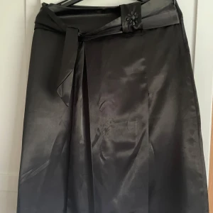 Svart kortkjol i satin från Puane  - Stilren svart långkjol i glansigt satinmaterial från Puane. Kjolen har ett brett midjeband med dekorativ blomdetalj och knytband. Perfekt för dig som vill ha en elegant och klassisk look med en twist.