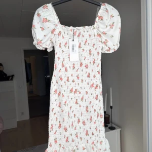 Vit blommig kortklänning med puffärm - Supersöt vit kortklänning från Chiquelle med rosa blommor, puffiga kortärmar och smockad överdel. Volang nertill ger extra söt vibe. Perfekt för sommarens alla tillfällen! 💐