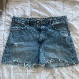 Blå jeansshorts från 157 - Snygga blå jeansshorts från 157 med råa, fransiga kanter och slitna detaljer både fram och bak. Klassisk femficksmodell med normal passform och dragkedja. Perfekta för sommaren och ger en avslappnad vibe.