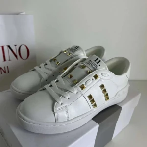 Valentino Rockstud vita sneakers - Snygga vita Valentino Rockstud sneakers i skinn med guldiga nitar och klassisk snörning. Låg modell med rund tå och platt sula. Ikoniska detaljer längs sidorna och hälen ger en lyxig och edgy vibe. Perfekta för dig som vill sticka ut med stil.