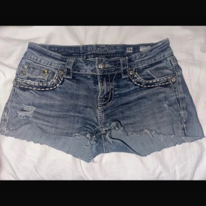 Miss Me jeansshorts med paljettdetaljer - Snygga blå jeansshorts från Miss Me med slitna detaljer och rå kant. Fickorna bak har stora peace-märken täckta av silvriga paljetter och coola kontrastsömmar. Perfekta för dig som vill sticka ut och gillar statement-detaljer.