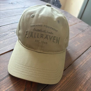 Beige keps från Fjällräven - Snygg beige keps från Fjällräven med broderad logga framtill och justerbar rem baktill. Klassisk böjd skärm och liten svensk flagga på sidan. Perfekt för dig som gillar outdoor och stilren design.