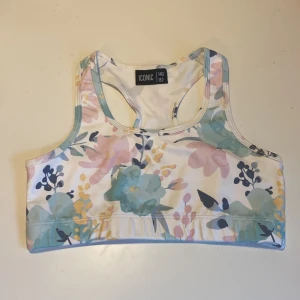 Sporttopp/bikini topp - Sportig och snygg topp från Iconic med blommigt mönster i pastelliga färger som ljusblått, rosa, gult och vitt. Modellen är ärmlös med brottarrygg och bred resår nedtill.