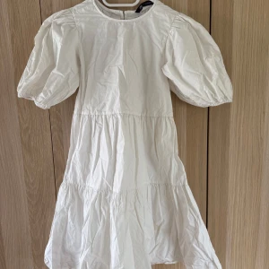 Vit kortklänning med puffärm från Zara - Supersöt vit kortklänning från Zara med puffiga kortärmar och volangkjol. Klänningen har öppen rygg med knapp och knyt bak, vilket ger en trendig och luftig känsla. Perfekt för varma dagar och snygg att styla med sneakers eller sandaler.