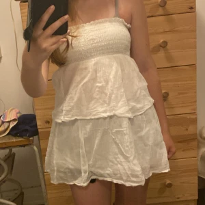 Vit mini klänning från Gina Tricot - Supersöt vit kortklänning från Gina Tricot med smock upptill och volangig kjol. Klänningen är axelbandslös och har en luftig känsla, perfekt för varma dagar. Tillverkad i ett lätt och skönt bomullstyg.