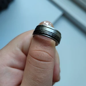 Bred silverfärgad ring med grekiskt mönster - Cool bred ring i silverfärg med ett svart, inristat grekiskt mönster runt hela ringen. Ringen har en matt finish och känns både stilren och edgy. Mönsterna på ringen kan snurra. 