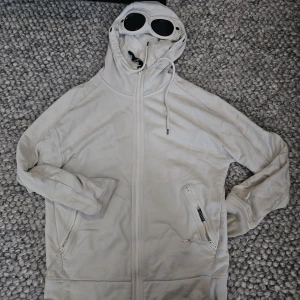 C.P Company - Säljer en ikonisk vit hoodiejacka från C.P. Company med inbyggda goggles i huvan. Jackan har dragkedja framtill, två fickor med dragkedja och tumhål i muddarna. Perfekt för dig som vill sticka ut med streetwear-vibe. Passar M oxh s skir mycke. Kostar ny 2500lr mitt pris 445kr