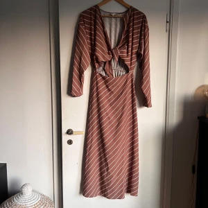 Randig långklänning med knyt och cut-out - Snygg långklänning i brun med vita ränder, v-ringad med knyt framtill och cool cut-out vid midjan. Långa ärmar och elastisk detalj i midjan för skön passform. Perfekt för dig som gillar en trendig och avslappnad stil. Obs defekt syns på de två sista bilderna 🌸