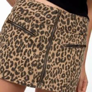 Leopardmönstrad kortkjol med dragkedja - Snygg kortkjol i leopardmönster med prislappen kvar och coola dragkedjedetaljer framtill och på fickorna. Kjolen är beige med svarta fläckar och har en tight passform som sitter snyggt på kroppen. Perfekt för dig som vill sticka ut med en trendig look.