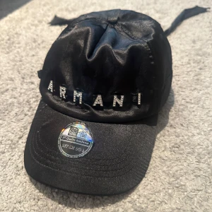 Svart Armani keps med strassdetaljer - Svart keps från Armani i glansigt material med stora bokstäver i strass framtill. Kepsen har knytband baktill och klassisk böjd skärm.
