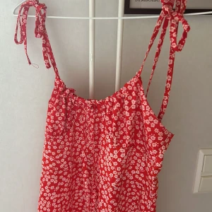 Röd blommig sommarklänning med knyt - Supersöt röd kortklänning med vita blommor och smala axelband som knyts upptill. Lätt och luftig modell, perfekt för varma dagar. Klänningen har en avslappnad passform och är tillverkad i ett mjukt tyg som känns skönt mot huden.