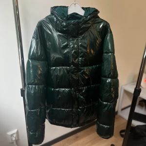 Mörkgrön pufferjacka - Zara (Oversized) - Säljer en riktigt snygg mörkgrön pufferjacka med glansig finish och huva. Jackan är vadderad, har långa ärmar och stängs med både dragkedja och knappar framtill. Perfekt för kalla dagar när du vill hålla stilen och värmen.