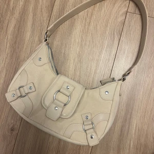 Beige axelväska med silvriga detaljer - Snygg beige axelväska i mockaimitation med silvriga metalldetaljer och dragkedja. Väskan har en kort axelrem, dekorativa spännen och en ficka framtill med magnetlås. Perfekt storlek för det viktigaste och ger en clean look till din outfit.