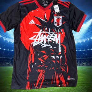Japan x Stüssy x Adidas fotbollströja M - Unik svart och röd Japan fotbollströja från Adidas i samarbete med Stüssy. Framsidan har en stor grafisk anime-inspirerad illustration och Japan-emblem på bröstet. Röd V-ringad krage och ärmslut, samt röda Adidas-ränder på axlarna. Tillverkad i lätt och ventilerande Aeroready-material. Storlek M,S
