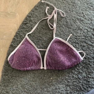 Glittrig lila bikiniöverdel Becksöndergaard - Triangelformad bikiniöverdel i glittrigt lila tyg med vita kanter och knyt i nacken och ryggen. Perfekt för strandhäng och sommar vibes. Snygg och trendig modell som verkligen sticker ut.