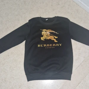 Svart Burberry sweatshirt med guldtryck - Svart sweatshirt från Burberry med stort guldtryck framtill som visar en riddare på häst och texten 'BURBERRY LONDON'. Tröjan har rund halsringning och långa ärmar. Perfekt för dig som vill ha en stilren men ändå lyxig look.