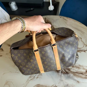 Louis Vuitton keepall 50  - Säljer en klassisk Louis Vuitton Keepall 50 resväska i brunt med ikoniskt monogrammönster och detaljer i ljust skinn. Väskan har två robusta handtag, dragkedja och snygga guldfärgade metalldetaljer. Perfekt för dig som vill resa med stil och visa upp en tidlös design.
