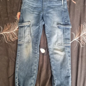 Blå cargopants från H&M, stl 134 - Snygga blå cargopants från H&M i jeansmaterial med stora fickor på sidorna och resår i midjan. Byxorna har en avslappnad passform och coola detaljer som ger en streetig vibe. Perfekta för dig som gillar praktiska och trendiga byxor.