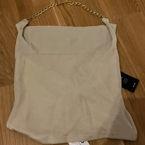 Beige glittrig linnetopp med kedjedetalj - Supersnygg beige linnetopp från NA-KD med glittrigt tyg och en cool guldfärgad kedja som axelband. Toppen har en draperad waterfall-ringning som ger en trendig look. Perfekt att styla med jeans eller kjol för en extra touch.