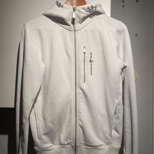 Vit hoodiejacka från Sail Racing - Vit hoodiejacka från Sail Racing med dragkedja framtill, bröstficka med dragkedja och logga på bröstet. Jackan har huva med snörning och två sidofickor. 