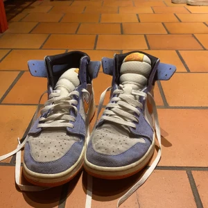 Nike Jordan 1 i blått och orange mocka - Nike Jordan 1 sneakers med högt skaft i ljusblå och grå mocka, orange detaljer och vit sula. Swoosh-logga i orange på sidan, vita skosnören och svart innerfoder. Sulan har coolt mönster i lila och orange. Perfekta för dig som vill sticka ut med färg. Lite smutsiga men annars i helt perfekt skick👌