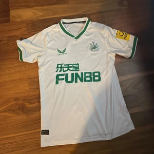 Newcastle United bortatröja 22/23 - Snygg Newcastle United bortatröja 22/23 med gröna detaljer. Kortärmad modell med grönt tryck och klubbmärke på bröstet, samt sponsorlogga. Kragen och ärmsluten har gröna ränder. Tillverkad i lätt och ventilerande material, perfekt för fotboll eller träning.