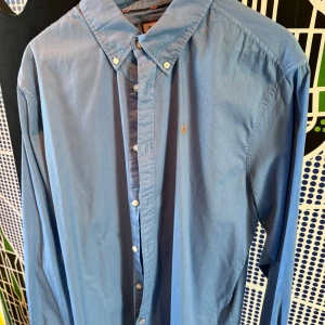 Blå skjorta från El Ganso - Snygg blå skjorta från El Ganso med klassisk button down-krage och broderad orange logga på bröstet. Skjortan har långa ärmar och vita knappar framtill. Tillverkad i mjuk bomull som känns skön mot huden. Perfekt för dig som gillar stilrena plagg med en twist.