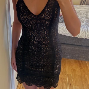 Svart spetsklänning med smala axelband - Supersnygg svart kortklänning med spets och smala axelband. Klänningen har v-ringning och figurnära passform, med ett vackert spetsmönster över hela plagget och nude-färgad underklänning. Perfekt för dig som vill sticka ut med en elegant look.