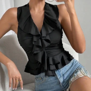 Svart volang topp med djup v-ringning - Supersnygg svart topp med volanger längs v-ringningen och nertill. Ärmlös modell som sitter figurnära och ger en feminin siluett. Perfekt att styla med jeans eller kjol för en trendig look. Säljer denna då den aldrig kommit till användning. Köpte från shein för 100kr.