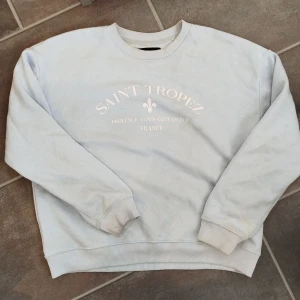 Ljusblå sweatshirt Saint Tropez - Ljusblå sweatshirt med vit text och tryck framtill där det står 'Saint Tropez Provence Alpes-Côte d'Azur France'. Tröjan har rund hals, ribbade muddar och är långärmad. Perfekt för en chill och avslappnad stil.