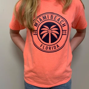 Säljer: Helt ny Miami Beach T-shirt – Oanvänd! 🌴☀️ - Snygg korallrosa t-shirt med stort tryck framtill där det står 'Miami Beach Florida' och en palm i mitten. T-shirten har rund hals, korta ärmar och är gjord i mjuk bomull. Perfekt för dig som gillar färg och vill sticka ut lite extra. Passar 12-13 åring. Lappen är kvar så helt ny! 