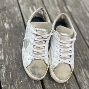 Vita sneakers från Philippe Model - Snygga vita sneakers från Philippe Model med beige mockadetaljer och silverfärgade inslag. Skorna har klassisk snörning, rund tå och platt sula. Ovandelen är i skinn och mocka, med en ikonisk sköld på sidan. Perfekta för en avslappnad streetstil. Defekter finns och det märks att de är använda!