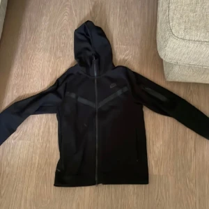 Svart Nike vindjacka med huva L - Svart vindjacka från Nike i storlek L. Jackan har hel dragkedja, huva och diskreta svarta detaljer på bröstet. Tillverkad i lätt och smidigt syntetmaterial, perfekt för aktiva dagar. Klassisk Nike-logga på bröstet och två sidofickor.