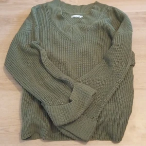 Olivgrön stickad tröja från STOCKH LM - Snygg olivgrön stickad tröja från STOCKH LM med v-ringad hals och breda ribbade muddar. Tröjan har lång ärm och en cool metallring med logga bak i nacken. Perfekt för lager-på-lager och chill höststil.