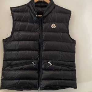 Svart dunväst från Moncler - Säljer en svart dunväst från Moncler med klassisk quiltad design och hög krage. Västen har två snedställda fickor med dragkedja och Moncler-logga på bröstet. Materialet är glansigt och lätt, perfekt för lager-på-lager-stil.