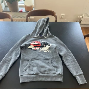 Grå Superdry hoodie med tryck - Snygg grå hoodie från Superdry med coolt japanskinspirerat tryck i rött, svart och vitt på bröstet. Tröjan har huva med vita snören och en stor magficka. Perfekt för vardagen.